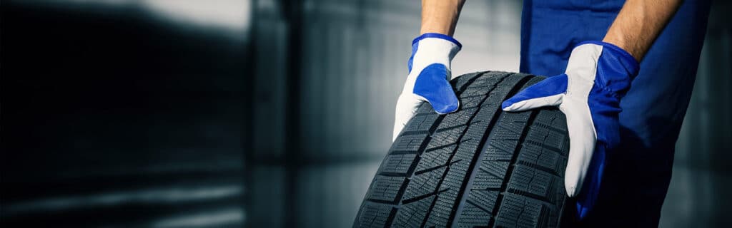 Changement de pneu hiver : Service rapide et professionnel Mains gantées (bleu et blanc) d'un technicien tenant un pneu hiver dans un garage. Préparation pour un changement de pneus.